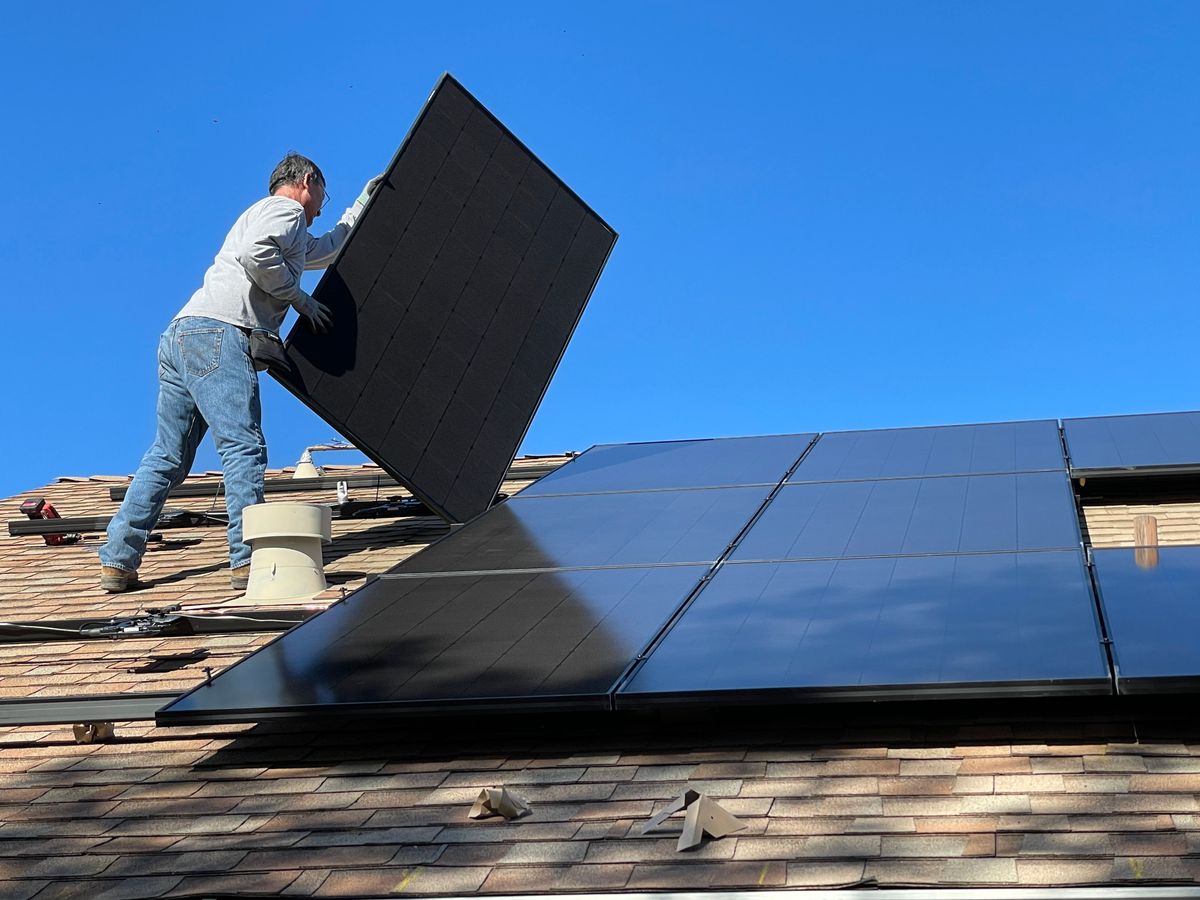 Installer carrying a black solar module across a composition shingle roof
